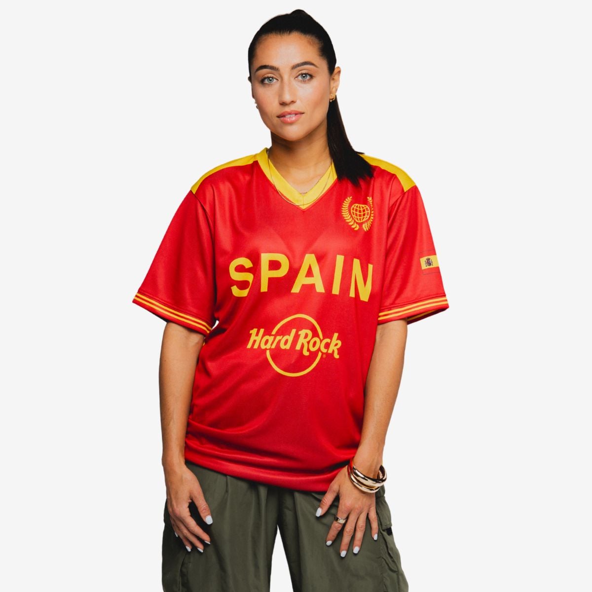 A person stands facing forward wearing a bright red Spain soccer jersey with yellow accents. The shirt prominently displays the word "SPAIN" across the chest and the Hard Rock text and circular graphic below. The individual has long hair and is dressed casually with green cargo pants and bracelets on one wrist. The background is plain white, keeping focus on the sports jersey and casual streetwear style.
